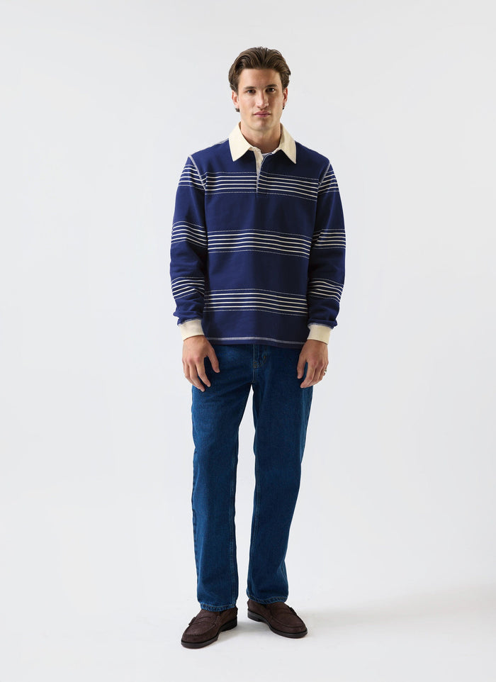 A model wears the Percival Editor Striped Rugby Polo Shirt in navy with dark blue jeans against a white studio background.