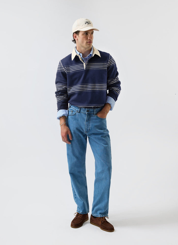 A model wears the Percival navy Editor Striped Rugby Polo shirt, styled with blue jeans, brown shoes and a cream cap.