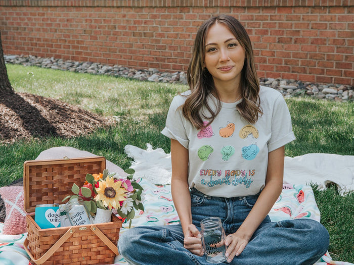 A young woman relaxes outdoors on a blanket beside a picnic basket, wearing the Sassy Woof "Every Body is a Beach Body Tee" with colorful animal graphics. Smiling at the camera, she holds a mason jar amid sunflowers and snacks.
