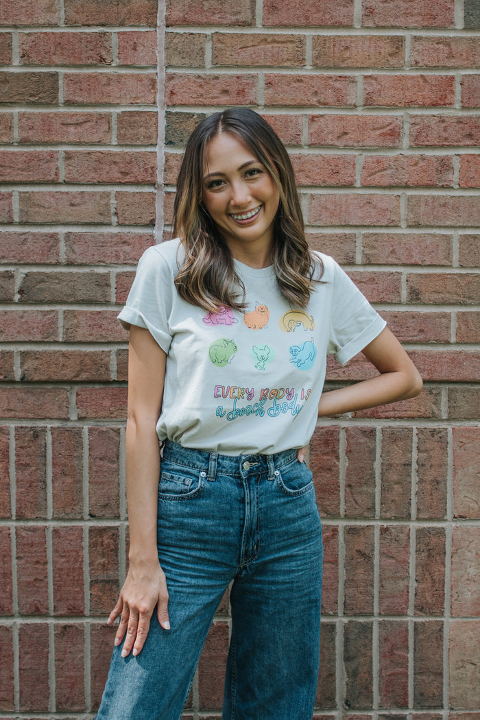 A woman with long brown hair smiles in front of a brick wall, wearing the Sassy Woof Every Body is a Beach Body Tee—a comfortable, lightweight shirt with colorful animal illustrations—and blue jeans.