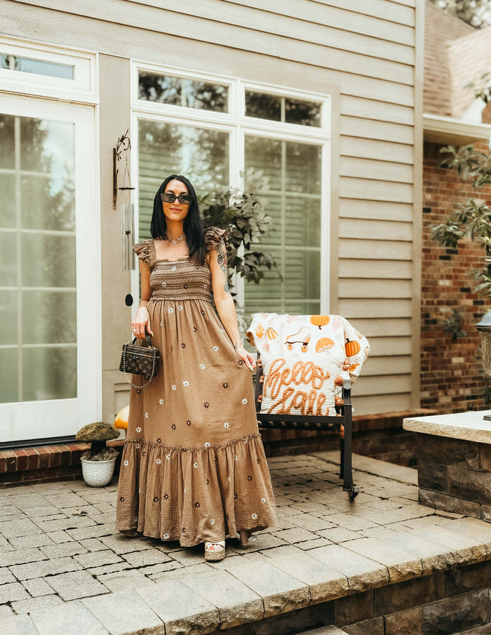 A woman stands on a stone patio in front of a house, wearing a long tan dress with embroidered details, holding a small patterned handbag. A bench with a 'Hello Fall' blanket is visible behind her.