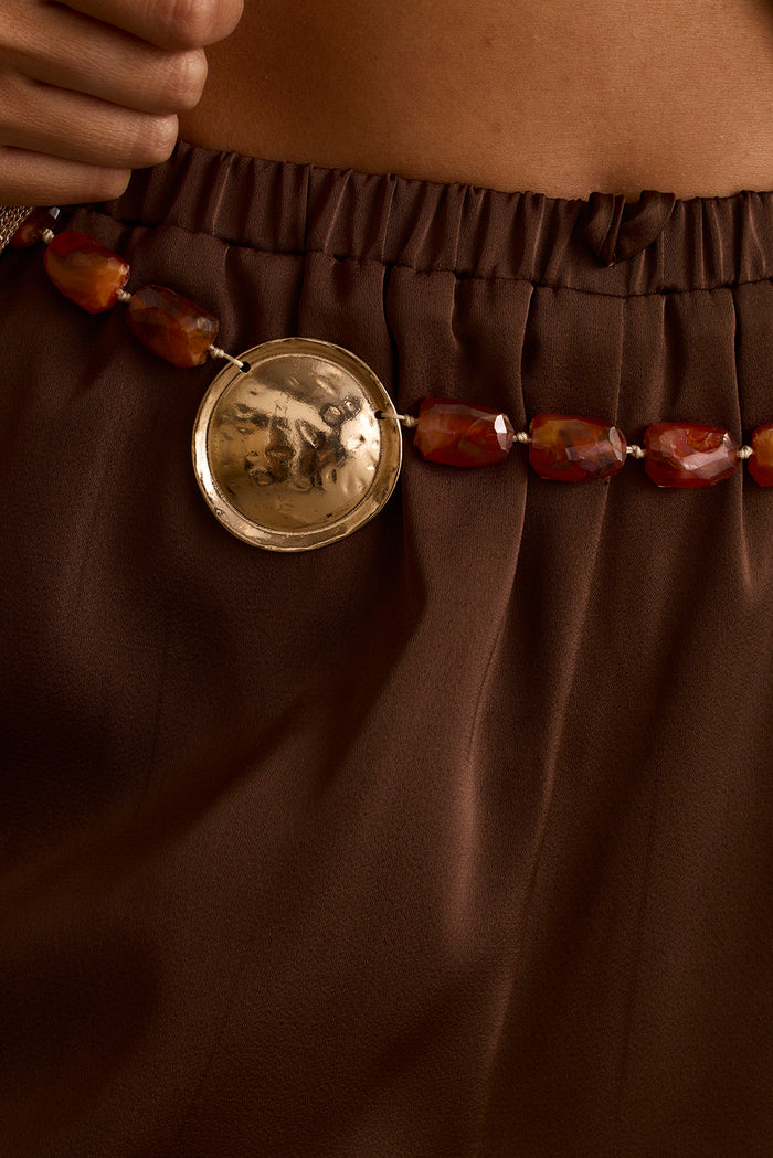 Close-up of hammered gold pendant and amber stone beads on a brown satin skirt.