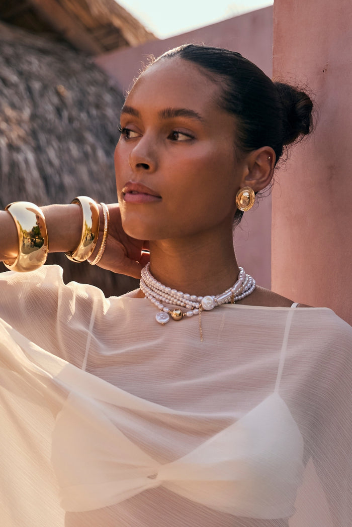 Textured gold coin stud earring worn with sheer ivory top and pearl chokers outdoors.