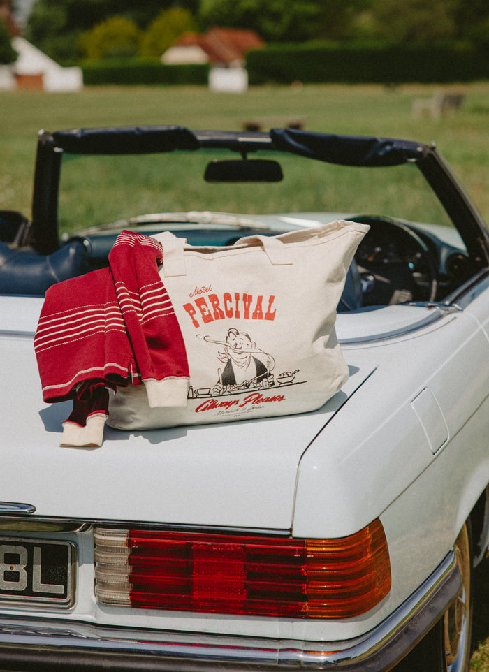 A Percival Gordon Large Tote in cream canvas with a red and black graphic print sits on the trunk of a white convertible car, next to a folded red striped knit sweater. The background is a blurred outdoor grassy field.