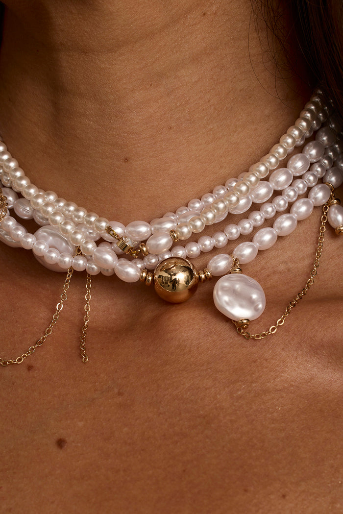 Layered pearl choker with gold ball pendant and baroque pearl drop, worn on neck.