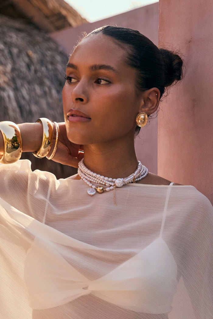 Layered pearl and gold choker worn on-model with sheer ivory top by a pink wall.