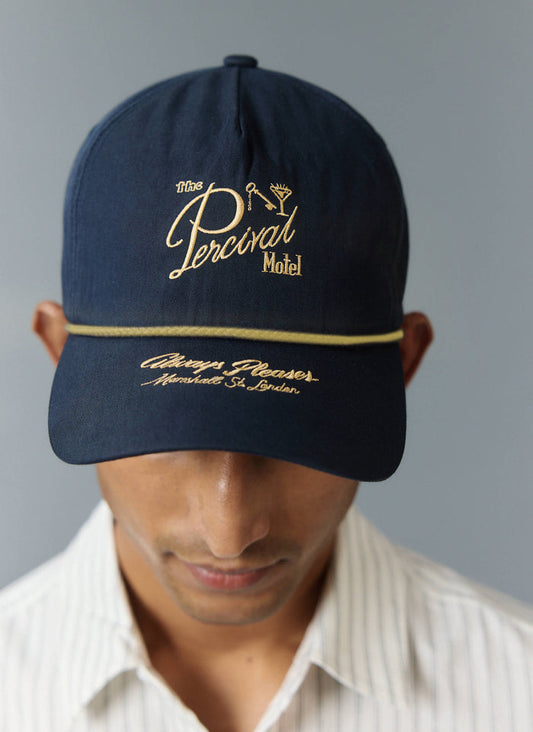 Percival Kingsley navy blue trucker cap with gold motel embroidery and rope, worn by a model in a white pinstripe shirt.