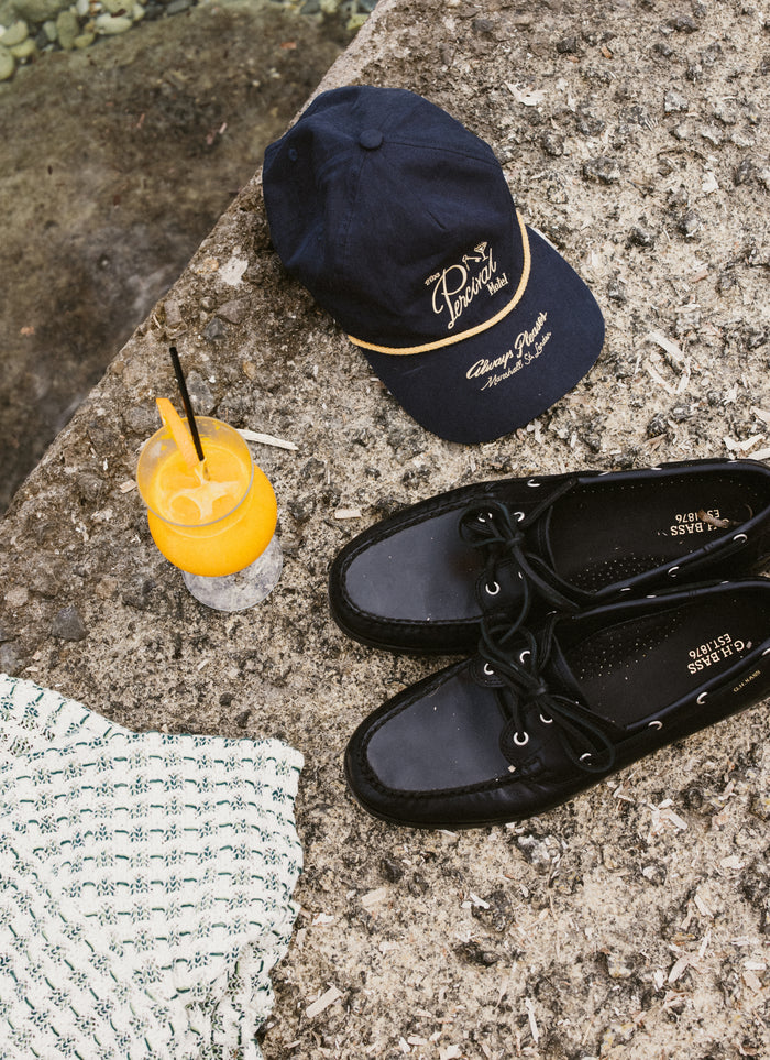Navy Percival Kingsley soft trucker cap with gold embroidery, styled on a stone ledge with boat shoes and a summer drink.
