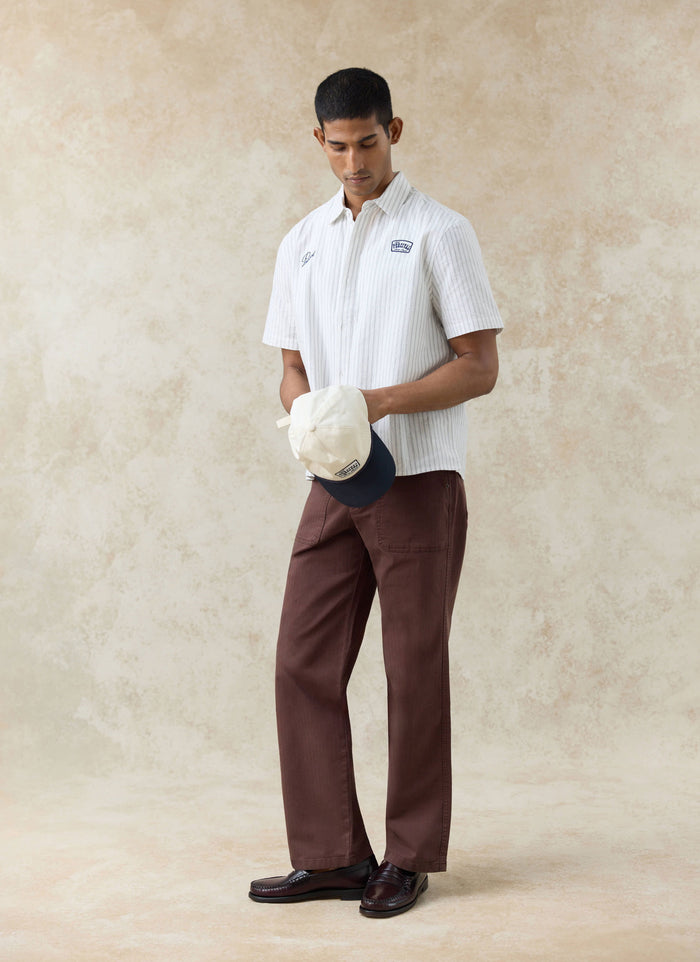 Percival Lucia Embroidered Shirt in cream stripe cotton, styled with brown trousers and a two-tone cap.