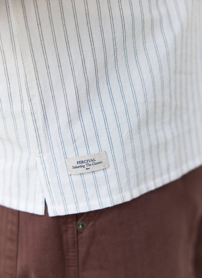 Close-up of the Lucia shirt in cream stripe cotton by Percival, highlighting the branded label at the bottom hem.
