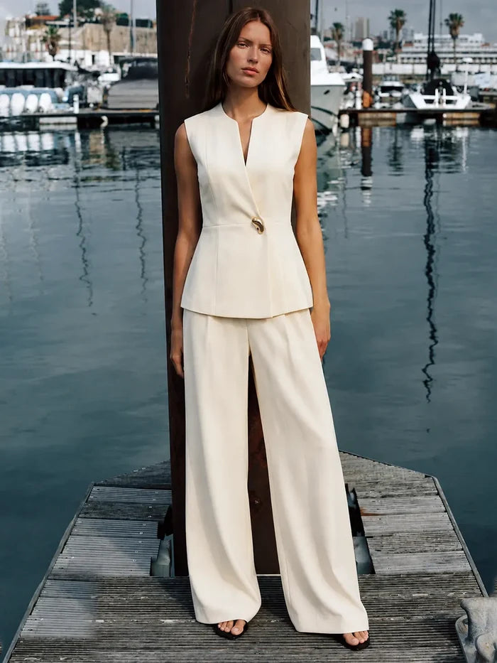 A woman stands on a wooden dock by the water wearing a sleeveless cream-colored two-piece suit with a structured peplum top and wide-leg trousers. The background shows a marina with boats and water.