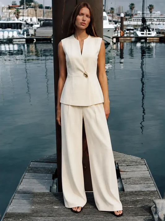 A woman stands on a wooden dock by the water wearing a sleeveless cream-colored two-piece suit with a structured peplum top and wide-leg trousers. The background shows a marina with boats and water.