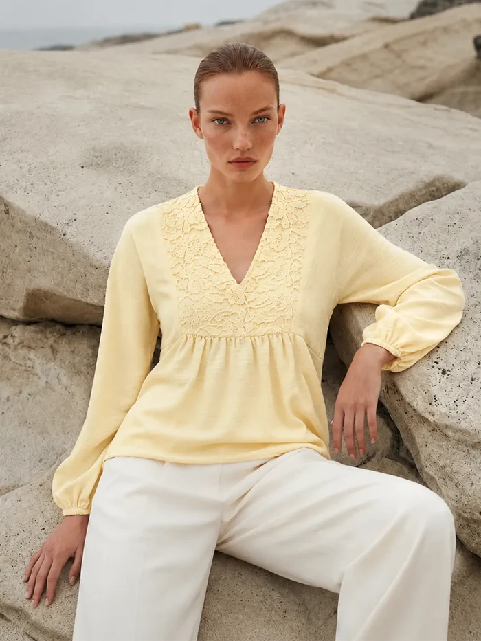 Woman wearing a yellow blouse and white pants sitting on rocks by the sea.