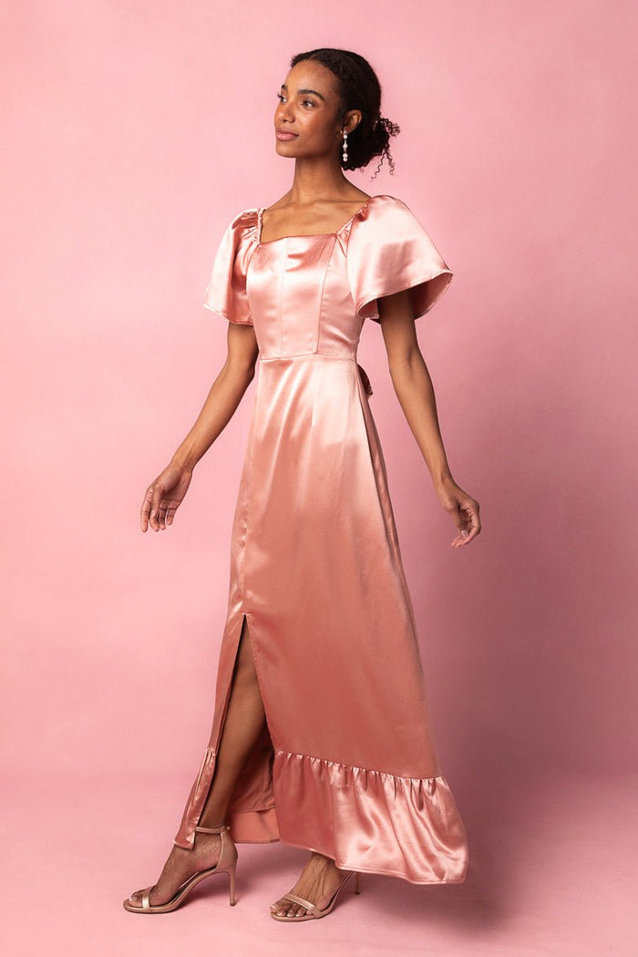 A woman stands against a matching pink background, looking to the side, wearing the Ivy City Co Maddie Dress in Apricot Crush Satin—a shiny, light pink short-sleeve dress featuring a square neckline and front slit. Styled with heels and dangling earrings, this look is perfect as a bridesmaid dress.