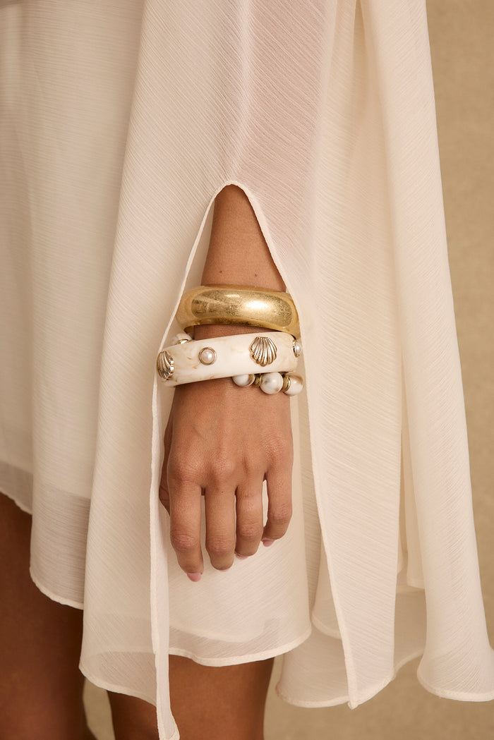 Stacked Mariners gold bangles with pearl and seashell accents on a wrist beside ivory chiffon.