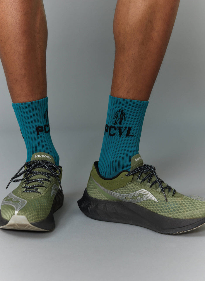 A model wearing the blue ribbed PCVL Sport Socks by Percival, showing the black jacquard graphic, styled with green trainers.