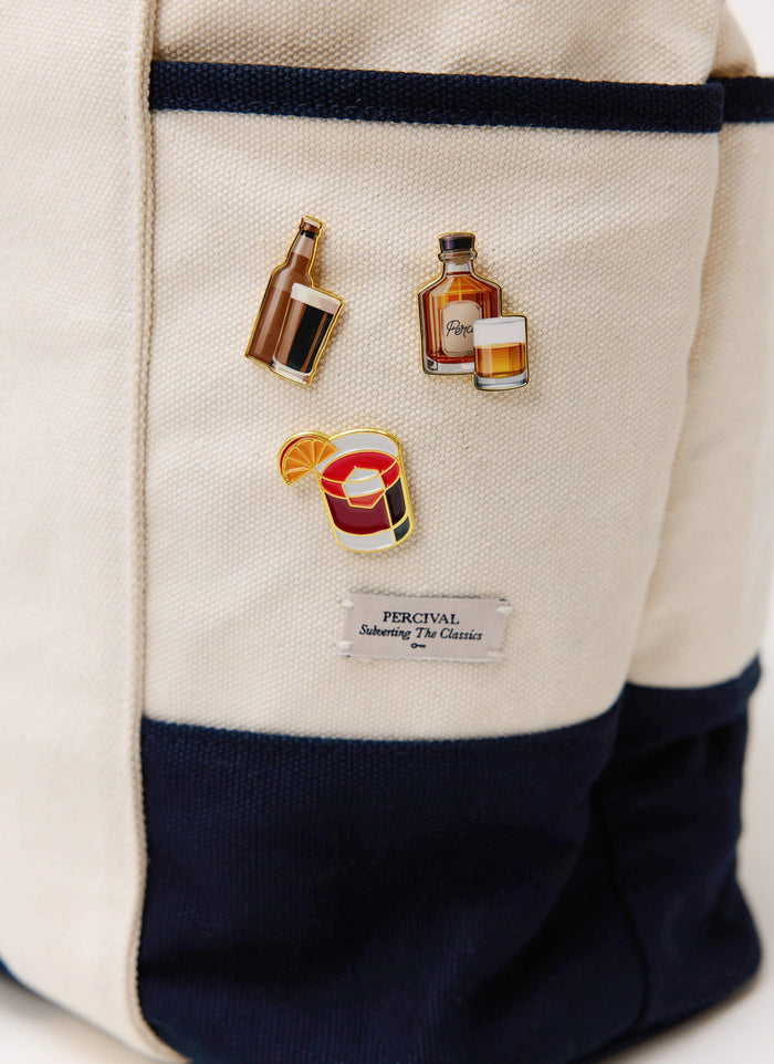A close-up of the Percival Negroni, whiskey, and beer pin badges pinned onto a cream and navy canvas tote bag.