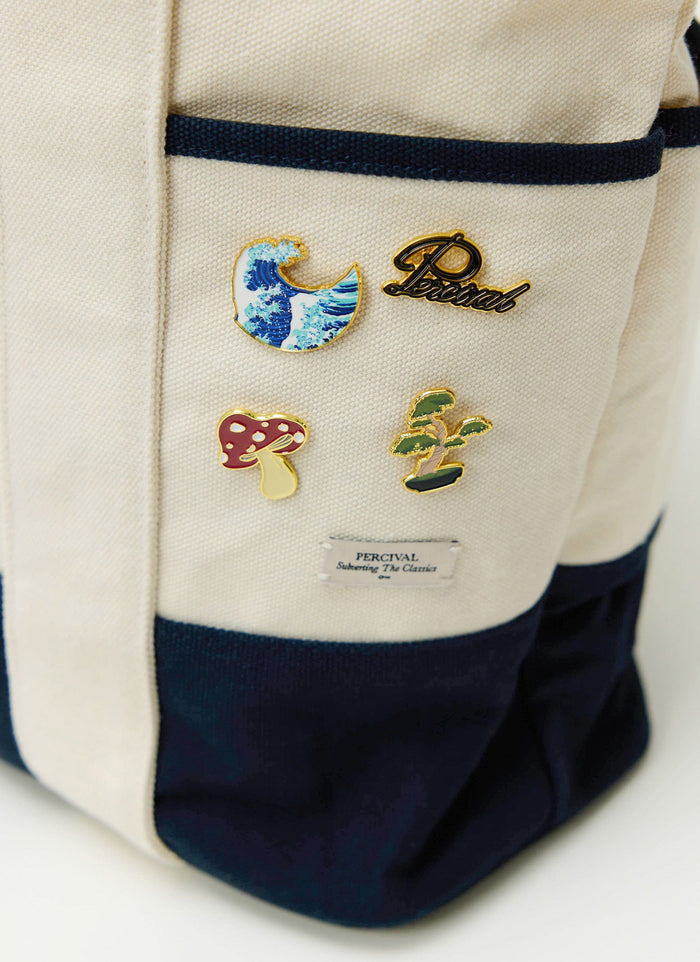 A collection of Percival pin badges, including the Wave pin, styled on a cream and navy canvas tote bag.