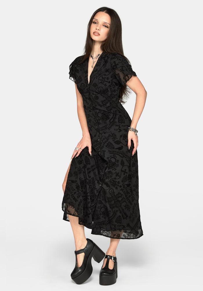A woman modeling a black midi dress with a sheer floral lace pattern, short flutter sleeves, and a wrap-style V-neckline, paired with black platform Mary Jane shoes against a light gray studio background.