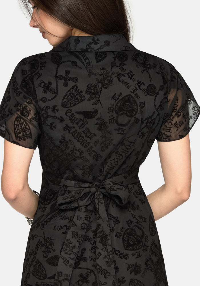 A woman shown from the back wearing a black short-sleeve dress with a sheer overlay and an allover gothic-style graphic print featuring skulls, crosses, and script.