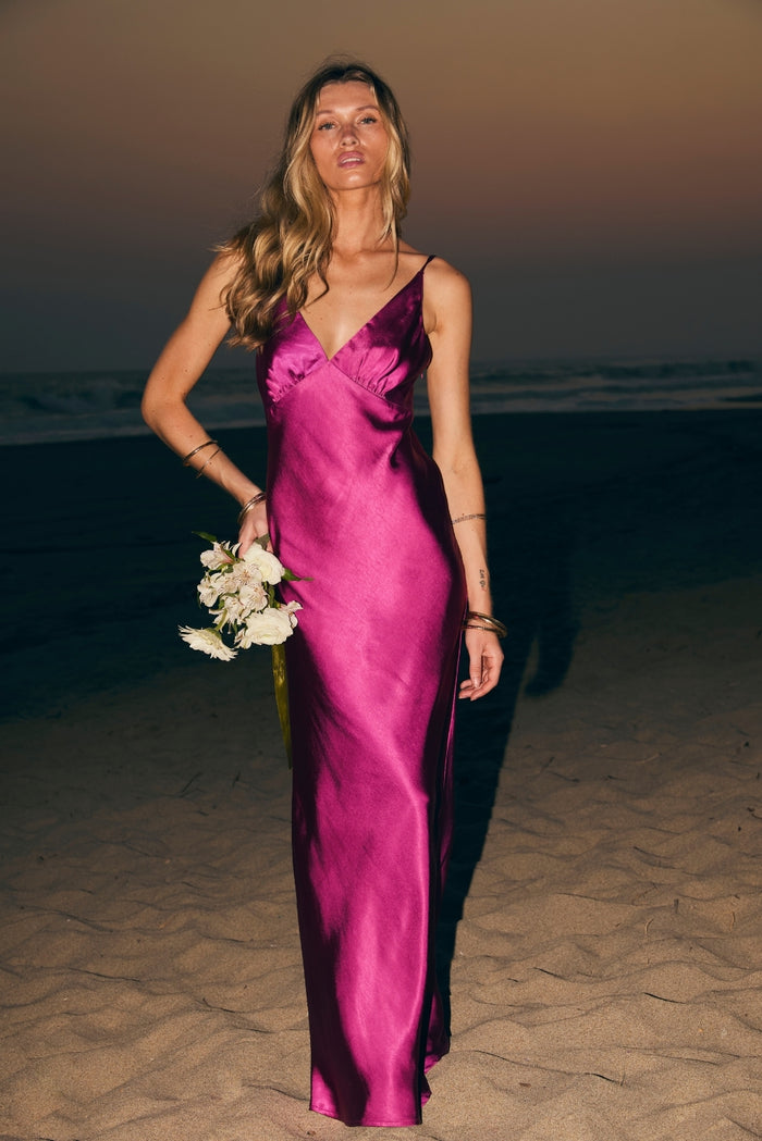Fuchsia satin maxi slip dress worn on a beach at dusk, holding white flowers.