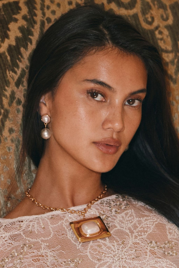 Close-up of a woman wearing gold pearl drop earrings and a gold pendant necklace with a beige lace top