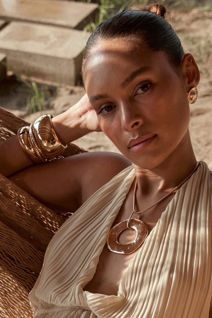 Stacked gold cuff bangles and organic pendant styled with a pleated cream resort dress.