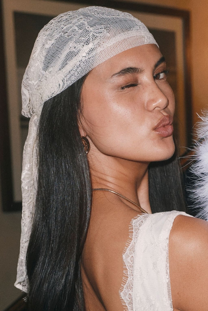Woman wearing a white lace scarf headwrap with a white lace-trim top and gold chain necklace