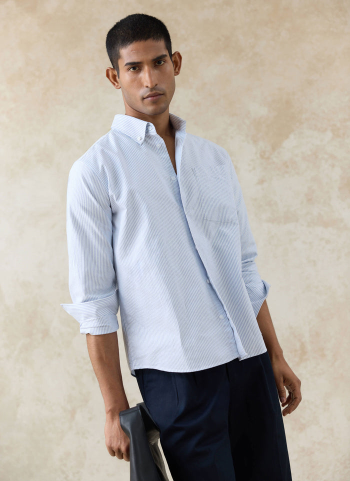 Model wearing a Percival blue pinstripe cotton Oxford shirt with rolled sleeves and navy trousers.