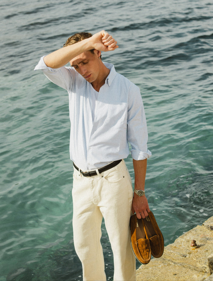 Men's blue striped cotton Oxford shirt by Percival, styled with cream trousers and worn by a model at the sea.