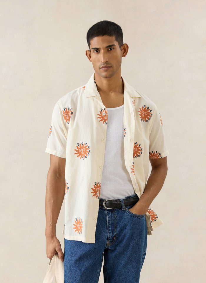 The Percival cream Sun Cuban shirt in cotton with orange sun embroidery, worn by a model with a white vest and jeans.