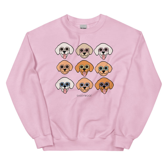 The "Goldens" sweatshirt by Sassy Woof is a light pink unisex crewneck with nine cartoon dog faces in colorful sunglasses, various fur colors, and "SASSY WOOF" printed below. Made from a comfy cotton blend.