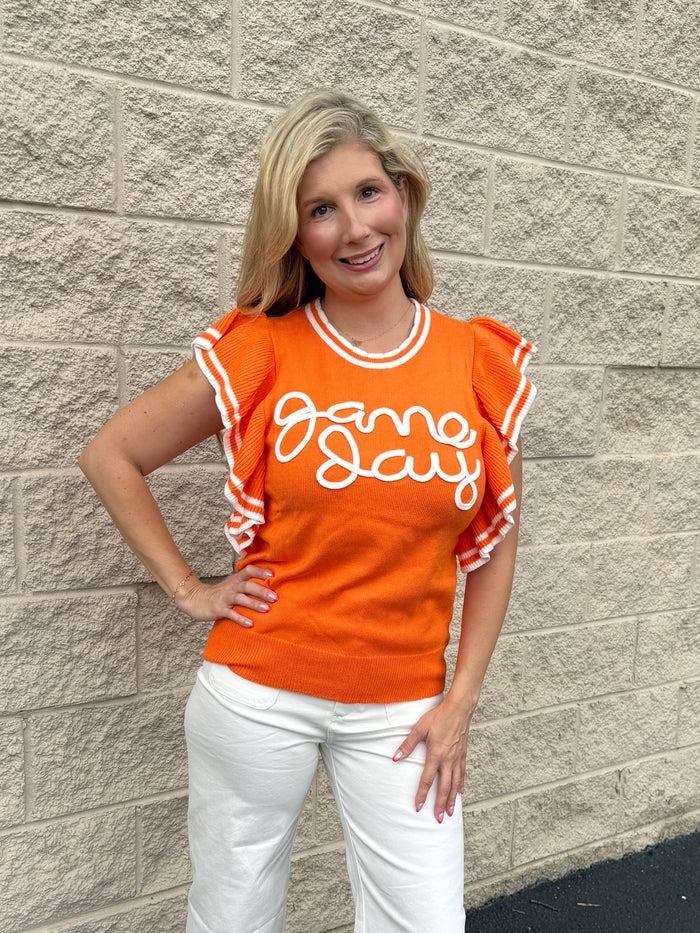 Woman wearing an orange 'Game Day' shirt and white pants standing against a stone wall.