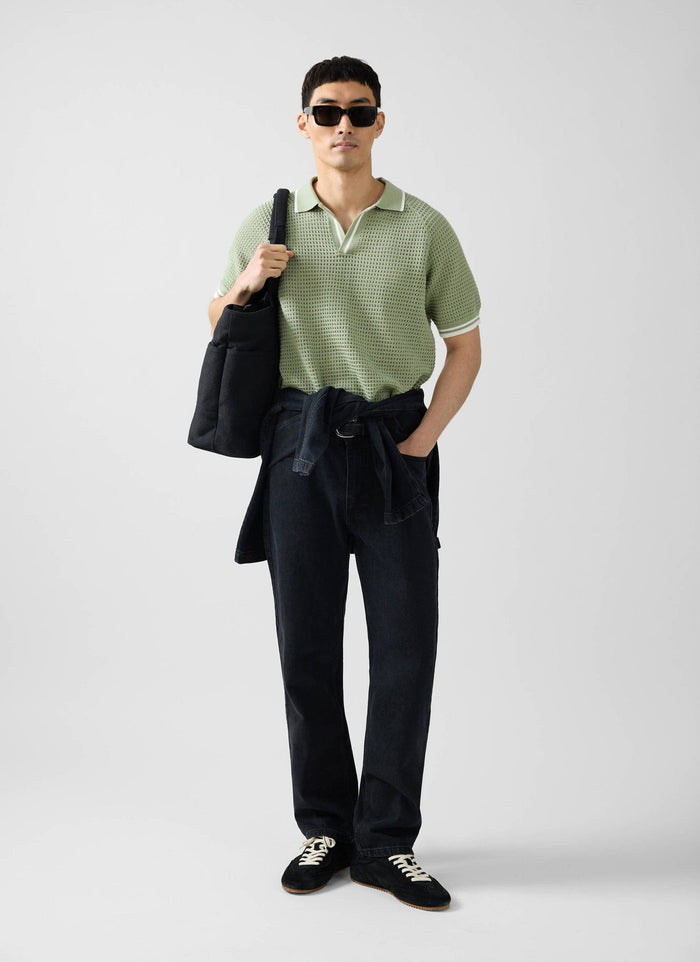A man wearing the Percival sage green Tennis Boxy Knitted Polo, black jeans, sunglasses, and carrying a black tote bag.