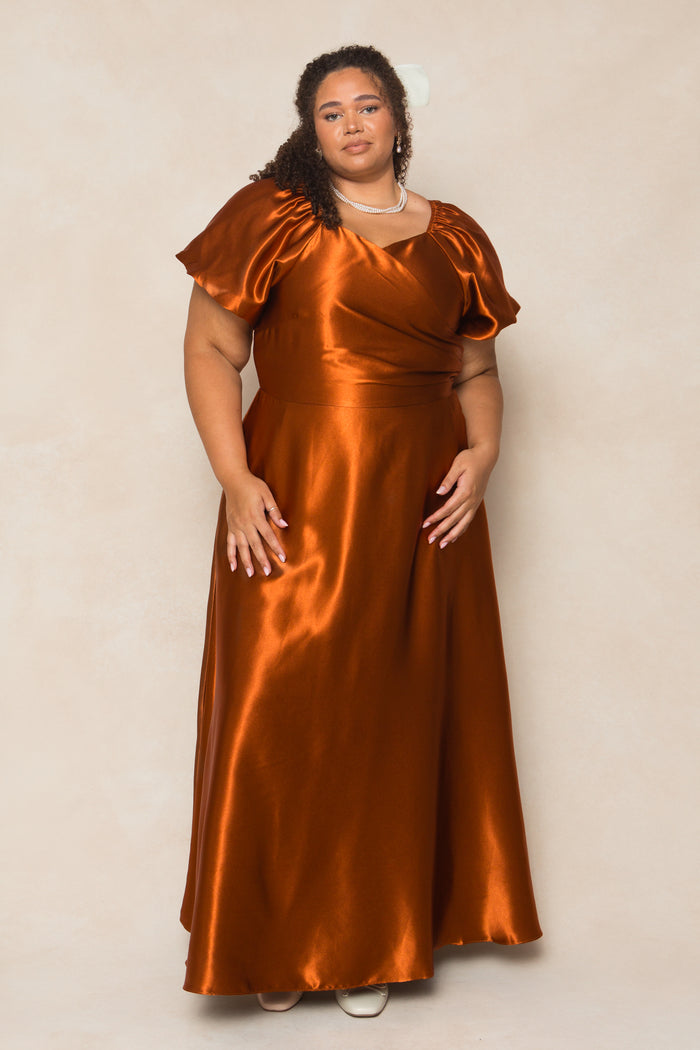 A woman in the Ivy City Co Tessie Dress in Amber Satin, a nursing-friendly, floor-length gown with short puff sleeves, stands against a light beige background. She has curly hair, wears a necklace, and white shoes peek out below.