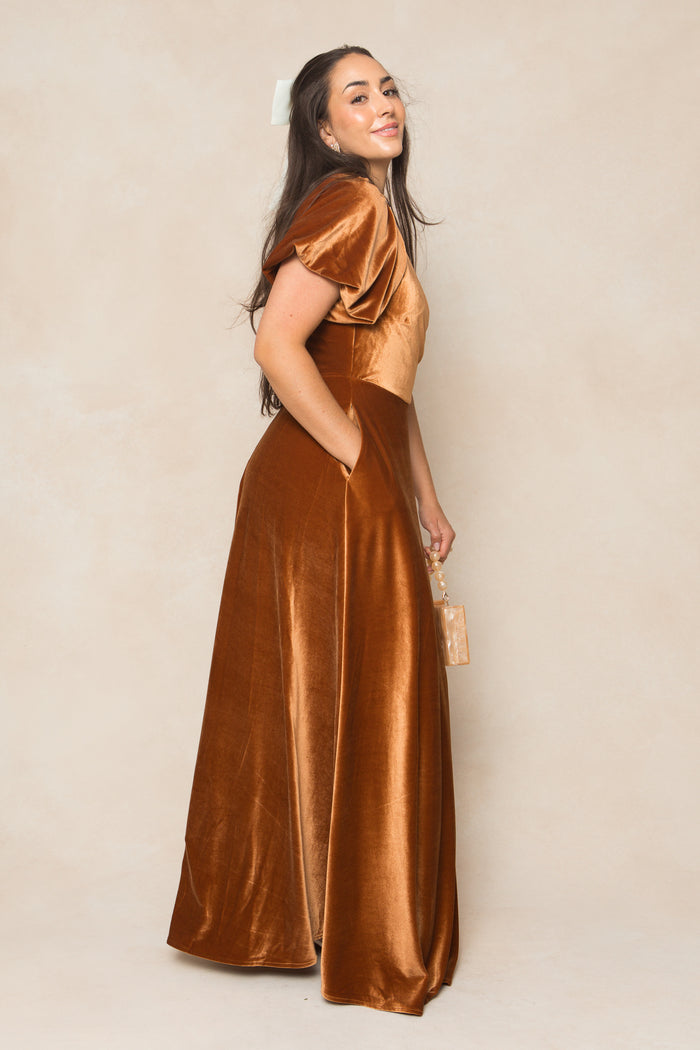 A woman smiles sideways in the Ivy City Co Tessie Dress in Amber Velvet, featuring short puff sleeves. She holds a small boxy handbag against a soft beige background.