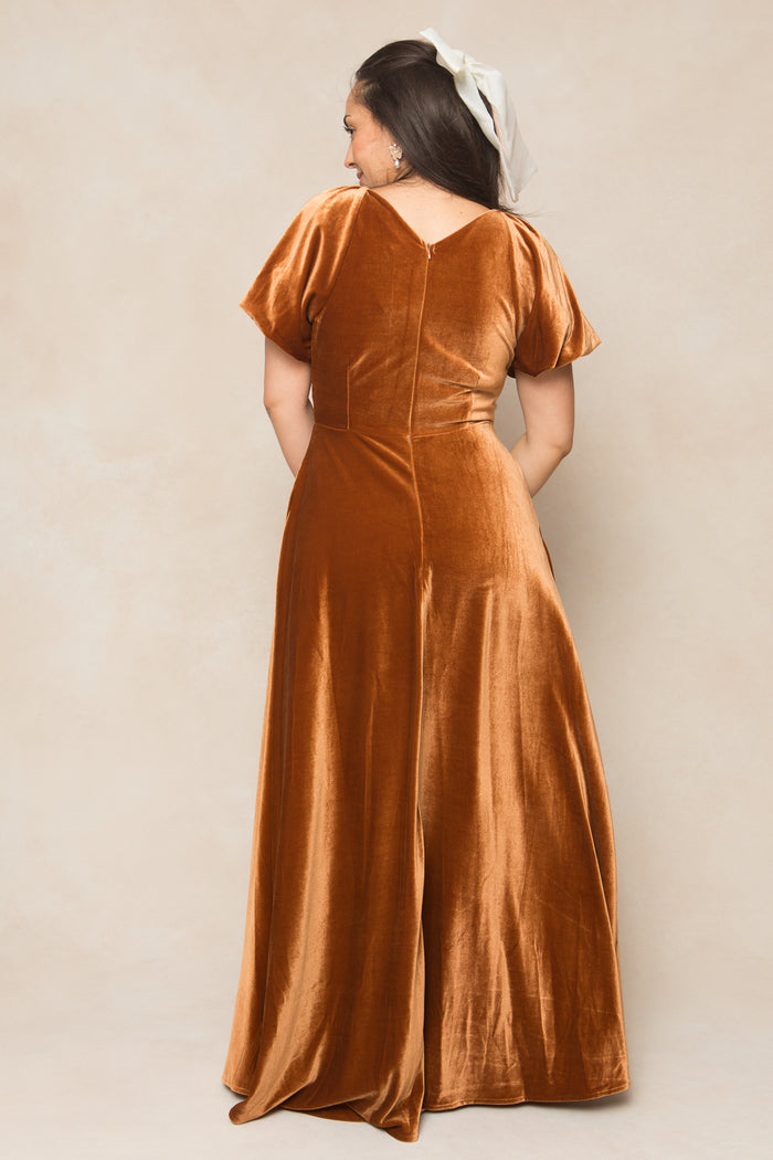 A woman in the Ivy City Co Tessie Dress in Amber Velvet, with long dark hair and a white bow, stands facing away with hands in pockets against a beige backdrop—a chic bridesmaids dress option.