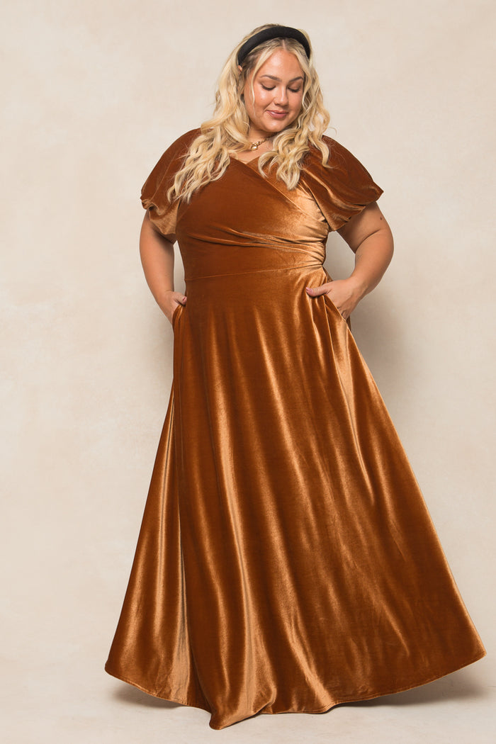 A woman with long blonde hair smiles with her hands in her pockets, wearing the Ivy City Co Tessie Dress in Amber Velvet—a stylish, floor-length bridesmaid dress featuring short sleeves and a timeless design against a beige background.