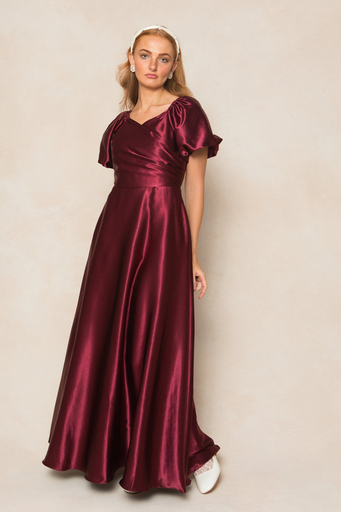 A woman wearing the Ivy City Co Tessie Dress in Wine Satin, featuring short puff sleeves and a fitted V-neck bodice, stands against a neutral background. She accessorizes with a headband and white shoes, perfect for fall bridesmaids.