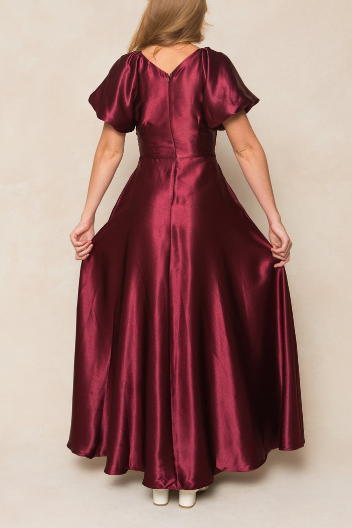 A woman faces away in the Ivy City Co Tessie Dress in Wine Satin, featuring short puff sleeves and a flared, floor-length skirt. She holds the sides of this elegant fall bridesmaid dress while white shoes peek out beneath the hem.